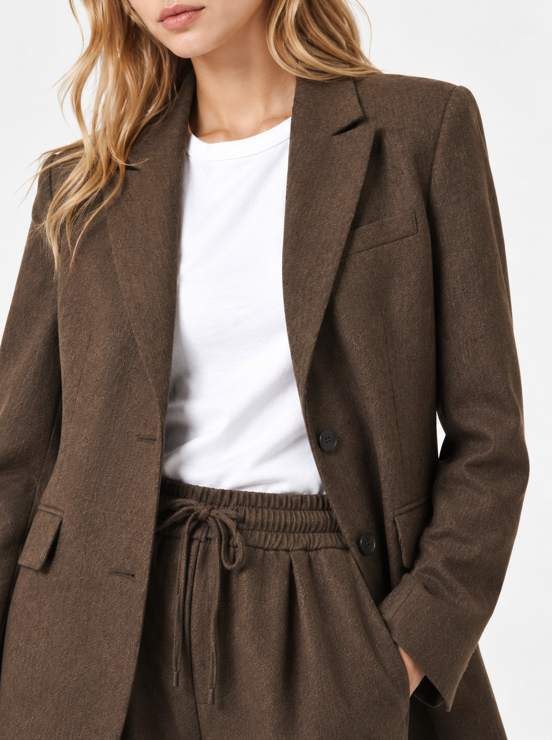 Eine Frau trägt einen braunen, taillierten Blazer und eine dazu passende Hose von Josy Braun sowie ein weißes T-Shirt. Das Outfit wirkt leger und dennoch professionell, und ihre linke Hand steckt in der Hosentasche. Ihr Gesicht ist teilweise außerhalb des Bildausschnitts. Das Set von Josy Braun wurde sorgfältig entworfen und in Portugal hergestellt.