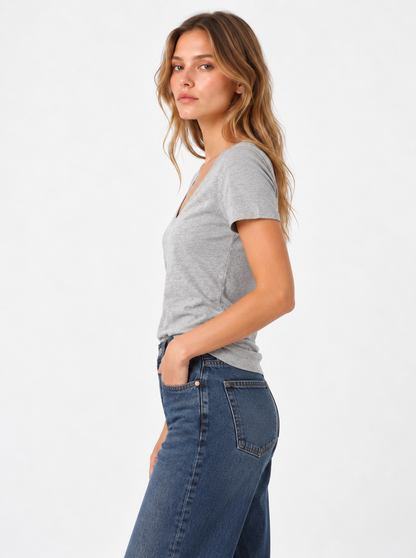 Eine Frau mit langen, welligen Haaren trägt ein graues T-Shirt mit V-Ausschnitt und eine blaue Jeans; sie steht im Profil, eine Hand in der Tasche, vor einem schlichten weißen Hintergrund.