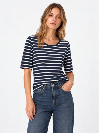 Eine Person, die ein T-Shirt in Marineblau mit weißen Streifen und eine blaue Jeans trägt, steht vor einem schlichten, hellen Hintergrund und hat eine Hand in der Hosentasche.