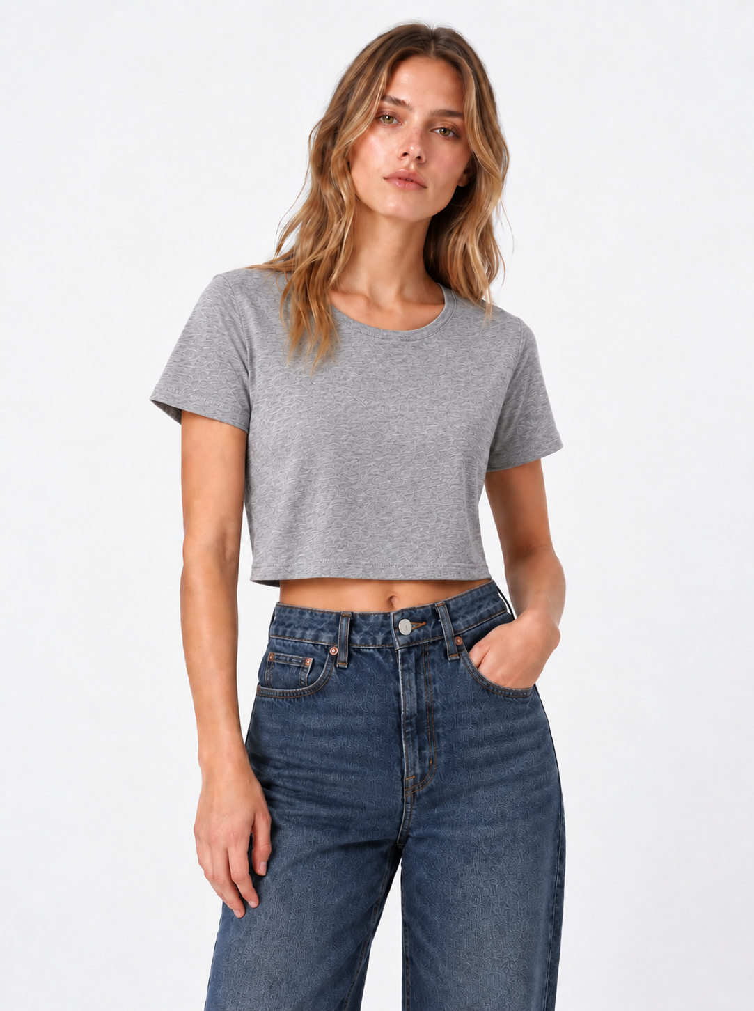 Eine Person steht vor einem schlichten Hintergrund und trägt ein in Portugal hergestelltes, kurz geschnittenes graues T-Shirt, dazu eine blaue Jeans mit hoher Taille und einen entspannten Gesichtsausdruck.