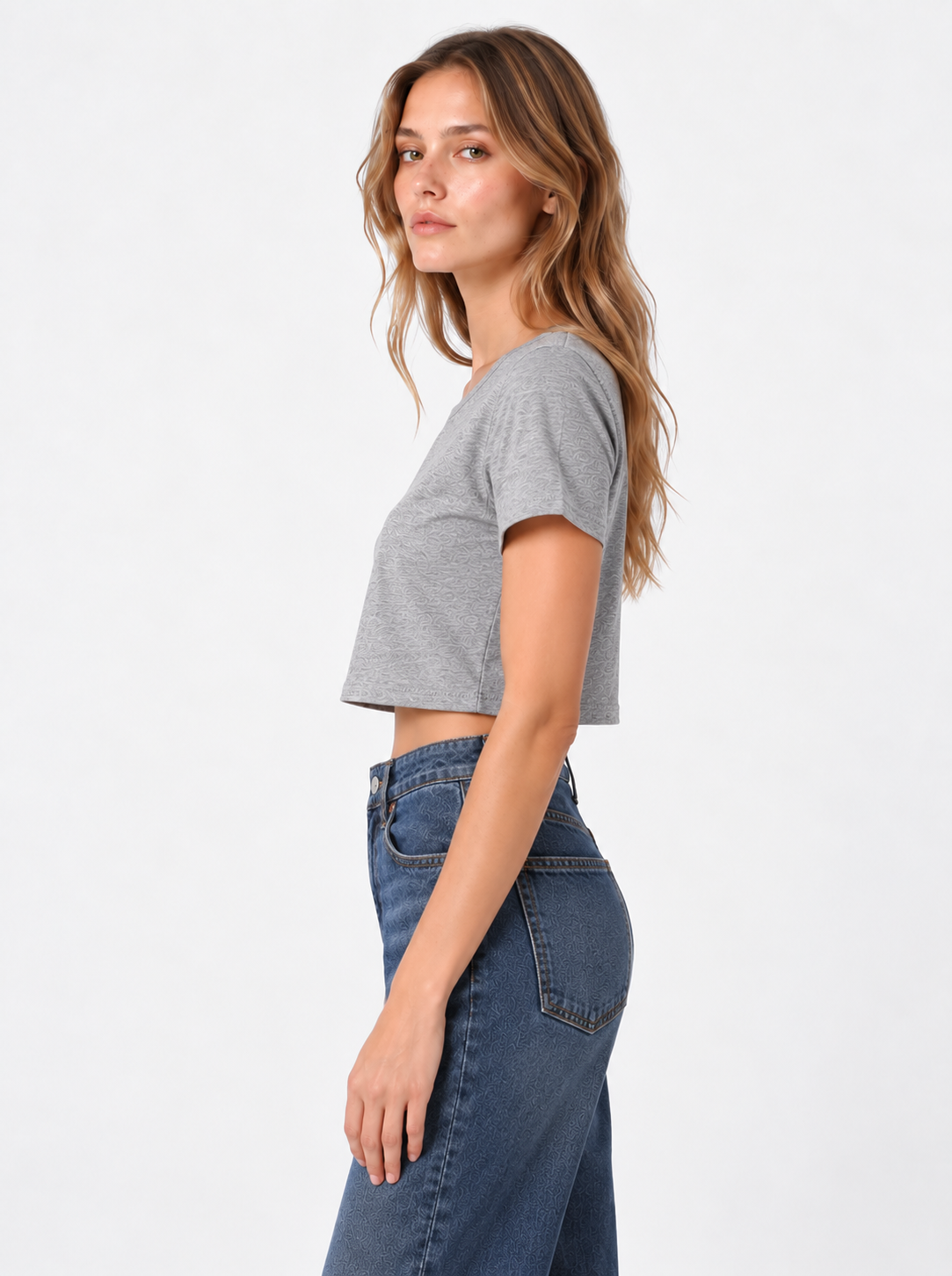 Eine Frau im Profil vor einem schlichten Hintergrund trägt das T-Shirt „Cropped Grau“ – hergestellt in Portugal aus Baumwolle – zu einer blau-hoch taillierten Jeans, hat langes, welliges Haar und einen neutralen Gesichtsausdruck.