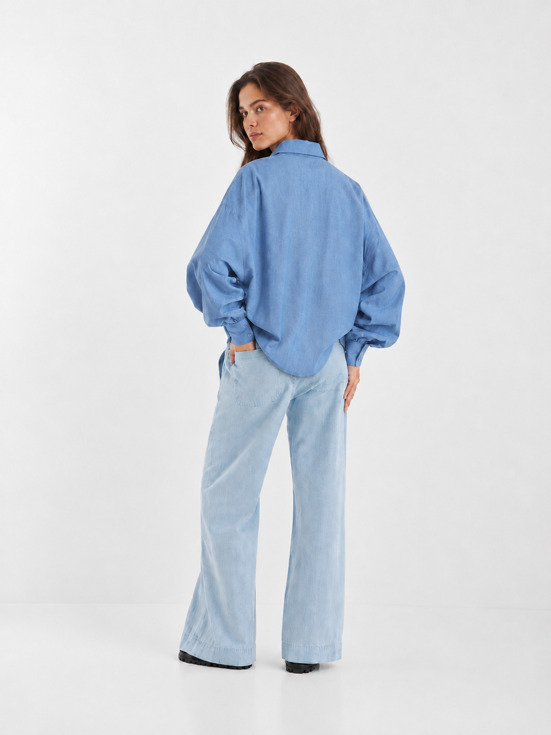 Eine Frau, von hinten gesehen, die eine hellblaue Bluse Antonia Slim Flanell Light Blue mit Knöpfen und voluminösen Ärmeln sowie eine hellgewaschene Jeans mit weitem Bein trägt.