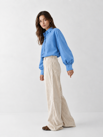 Eine Frau, die ein hellblaues Bluse Antonia Slim Flanell-Hemd mit Knöpfen trägt, das in eine cremefarbene Hose mit weitem Bein gesteckt ist.