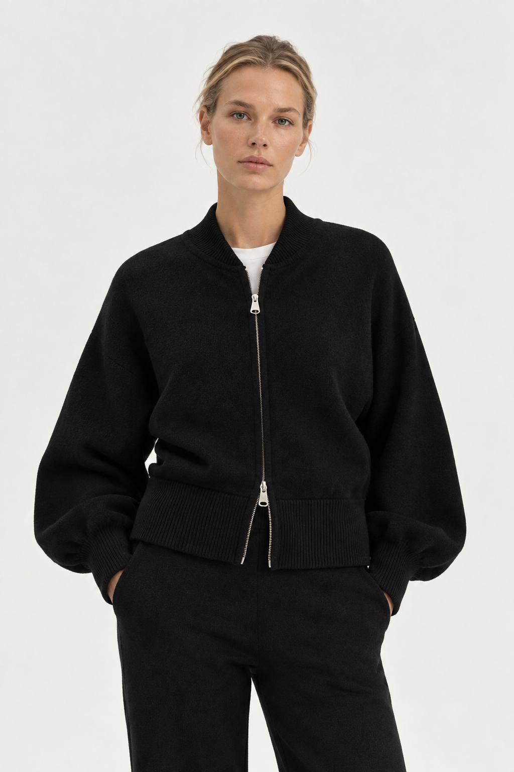 Eine Frau steht mit den Händen in den Taschen vor einem schlichten weißen Hintergrund und trägt den „Bomber Cardigan Schwarz“ aus weicher Merinowolle, kombiniert mit einer passenden Hose.