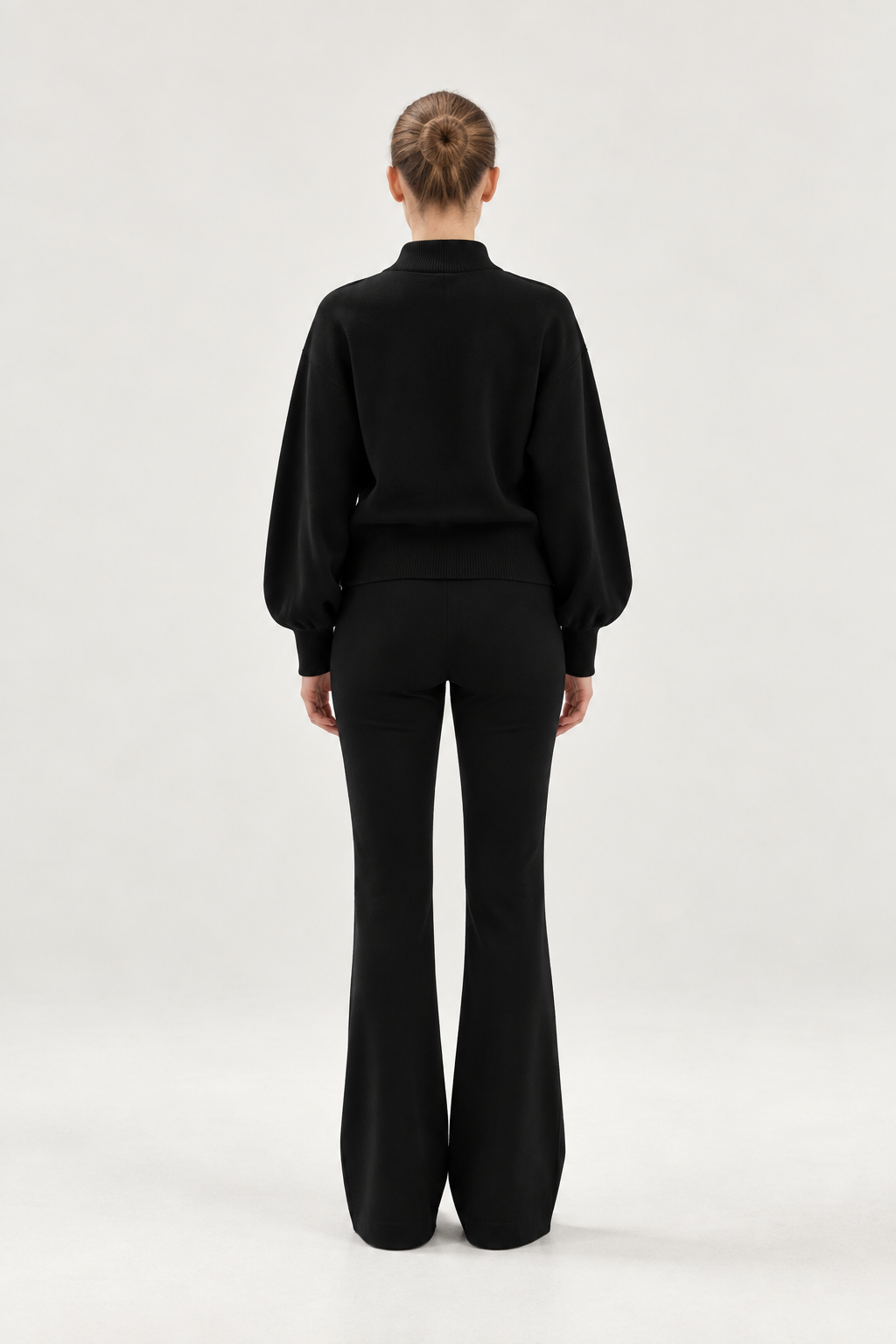 Eine Person steht mit dem Rücken zur Kamera und trägt ein schwarzes Langarmshirt sowie eine schwarze Schlaghose, kombiniert mit dem „Bomber Cardigan Schwarz“. Der aus weicher Merinowolle gefertigte Cardigan verleiht dem Outfit einen eleganten Touch. Die Haare sind ordentlich zu einem Dutt zusammengebunden, und der Hintergrund ist einfarbig und hell.