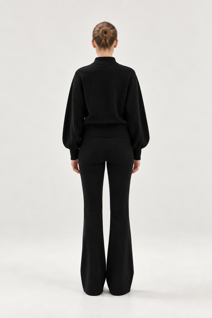 Eine Person steht mit dem Rücken zur Kamera und trägt ein schwarzes Langarmshirt sowie eine schwarze Schlaghose, kombiniert mit dem „Bomber Cardigan Schwarz“. Der aus weicher Merinowolle gefertigte Cardigan verleiht dem Outfit einen eleganten Touch. Die Haare sind ordentlich zu einem Dutt zusammengebunden, und der Hintergrund ist einfarbig und hell.