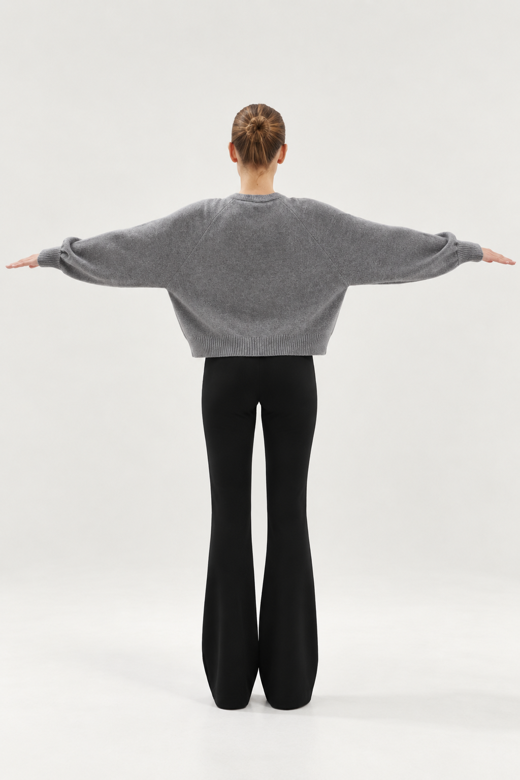 Eine Person steht mit ausgestreckten Armen da, von hinten gesehen. Sie trägt den „Antonia Short Cardigan Grau“ und eine schwarze Schlaghose, ihr Haar ist zu einem ordentlichen Dutt zusammengebunden, vor einem schlichten, hellen Hintergrund.