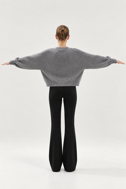 Eine Person steht mit ausgestreckten Armen da, von hinten gesehen. Sie trägt den „Antonia Short Cardigan Grau“ und eine schwarze Schlaghose, ihr Haar ist zu einem ordentlichen Dutt zusammengebunden, vor einem schlichten, hellen Hintergrund.