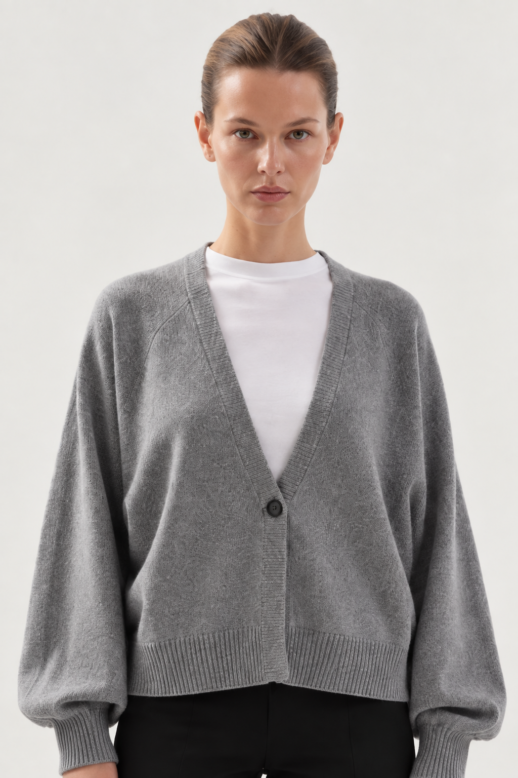 Eine Person, die ein weißes T-Shirt unter einer lockeren, grauen Antonia-Kurzstrickjacke mit einem einzigen Knopf trägt und vor einem schlichten, hellen Hintergrund steht.