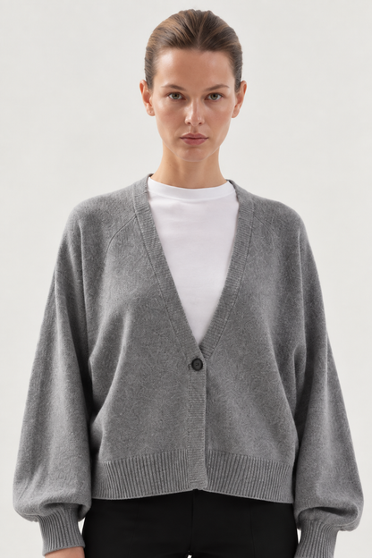 Eine Person, die ein weißes T-Shirt unter einer lockeren, grauen Antonia-Kurzstrickjacke mit einem einzigen Knopf trägt und vor einem schlichten, hellen Hintergrund steht.
