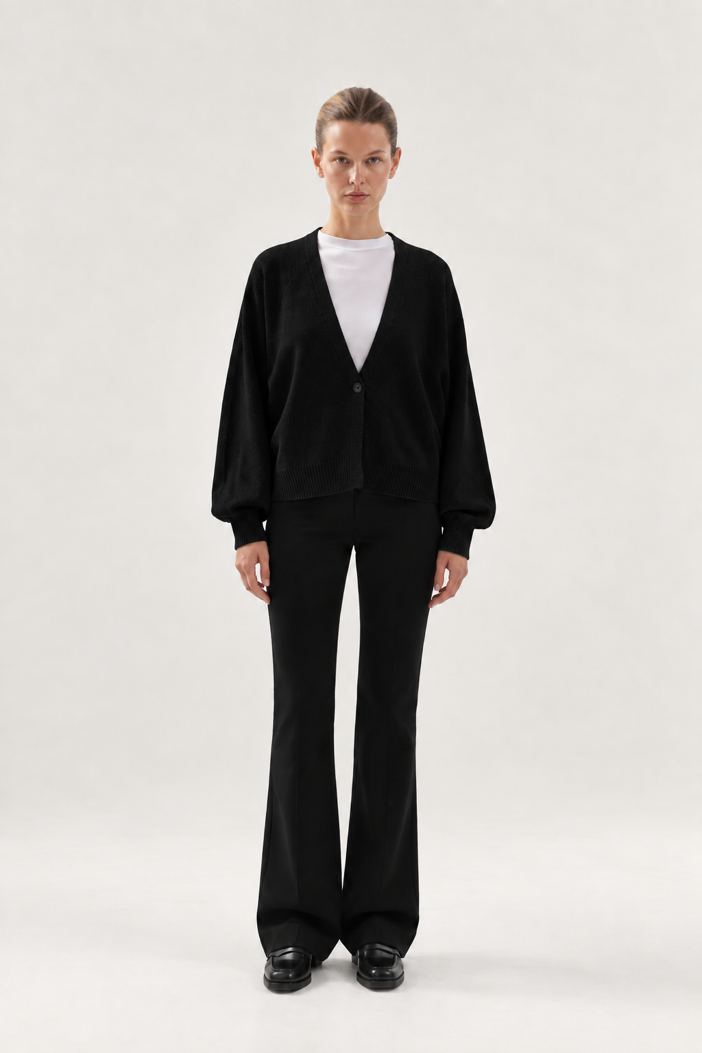 Eine Person steht mit dem Gesicht nach vorne vor einem schlichten, hellen Hintergrund und trägt den „Antonia Short Cardigan Schwarz“ aus Merinowolle, ein weißes T-Shirt, eine schwarze Schlaghose und schwarze Schuhe.