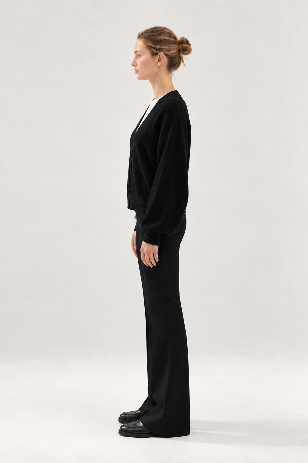 Eine Frau steht im Profil vor einem hellen Hintergrund. Sie trägt den Antonia Short Cardigan Schwarz, eine schwarze Hose und schwarze Schuhe, und ihr Haar ist zu einem ordentlichen Dutt zusammengebunden.