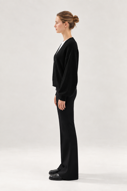 Eine Frau steht im Profil vor einem hellen Hintergrund. Sie trägt den Antonia Short Cardigan Schwarz, eine schwarze Hose und schwarze Schuhe, und ihr Haar ist zu einem ordentlichen Dutt zusammengebunden.