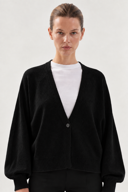 Eine Person, die den „Antonia Short Cardigan Schwarz“ über einem weißen Hemd trägt, steht mit dem Gesicht nach vorne vor einem schlichten, hellen Hintergrund.