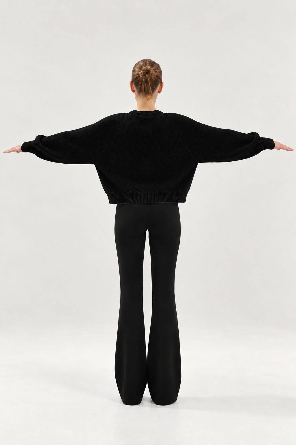 Eine Person steht mit ausgestreckten Armen mit dem Rücken zur Kamera und trägt den „Antonia Short Cardigan Schwarz“ sowie eine schwarze Schlaghose vor einem schlichten, hellen Hintergrund.