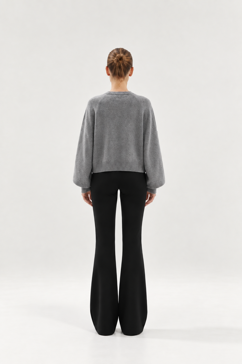 Eine Frau steht mit dem Rücken zur Kamera und trägt den „Antonia Short Cardigan Grau“ mit weiten Ärmeln sowie eine schwarze Schlaghose. Der Hintergrund ist einfarbig und hell.