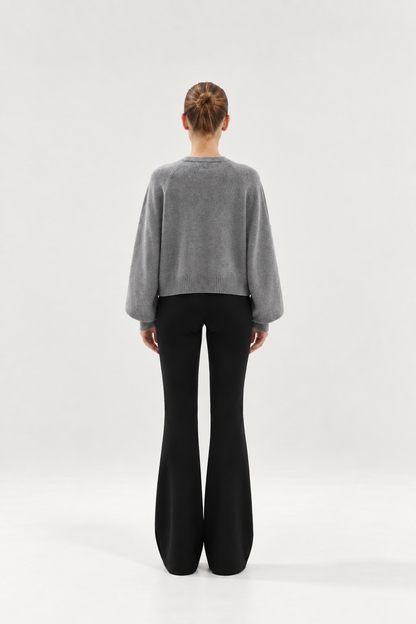 Eine Frau steht mit dem Rücken zur Kamera und trägt den „Antonia Short Cardigan Grau“ mit weiten Ärmeln sowie eine schwarze Schlaghose. Der Hintergrund ist einfarbig und hell.