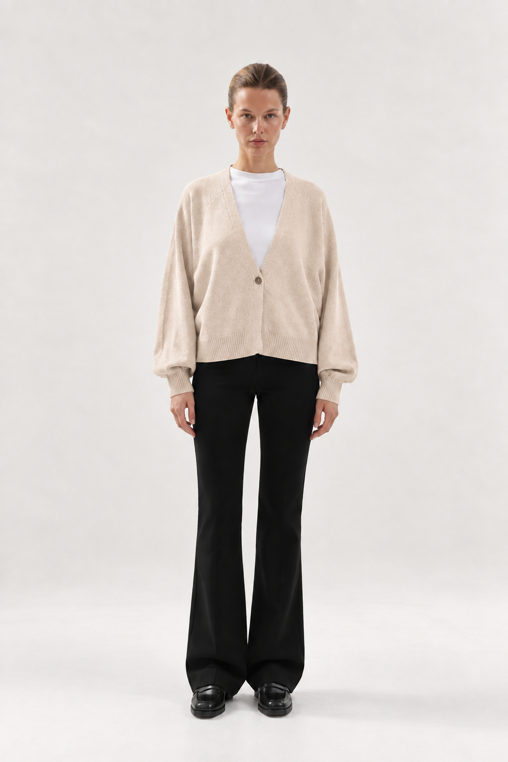 Eine Person trägt die Strickjacke Keeping It Casual, einen beigen Cardigan mit V-Ausschnitt und Knopfleiste über einem weißen T-Shirt, dazu eine schwarze Schlaghose und schwarze Loafer.