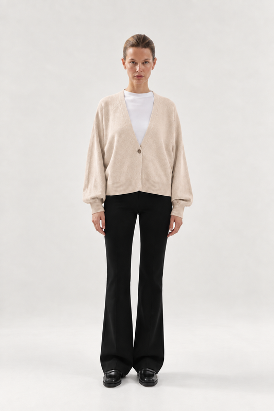 Eine Person trägt die Strickjacke Keeping It Casual, einen beigen Cardigan mit V-Ausschnitt und Knopfleiste über einem weißen T-Shirt, dazu eine schwarze Schlaghose und schwarze Loafer.
