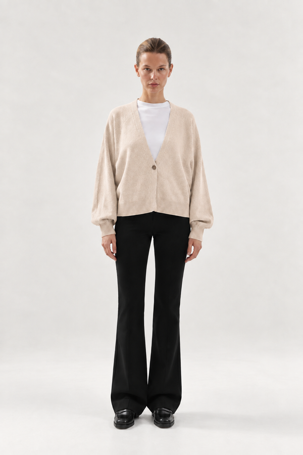 Eine Person trägt die Strickjacke Keeping It Casual, einen beigen Cardigan mit V-Ausschnitt und Knopfleiste über einem weißen T-Shirt, dazu eine schwarze Schlaghose und schwarze Loafer.