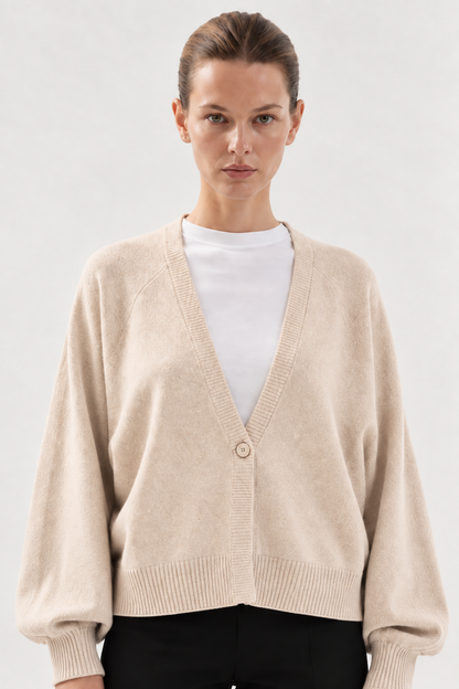 Eine Frau steht mit neutralem Gesichtsausdruck vor einem schlichten, hellen Hintergrund und trägt den „Antonia Short Cardigan Puder“ – eine beigefarbene Strickjacke aus Merinowolle mit einem einzigen Knopf –, die sie über einer weißen Bluse trägt.