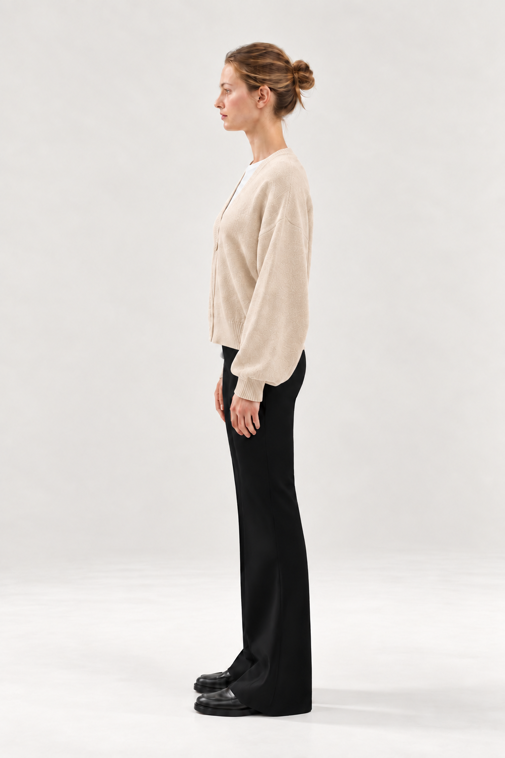 Eine Frau steht im Profil vor einem schlichten, hellen Hintergrund und trägt die beigefarbene kurze Strickjacke „Antonia Puder“, eine schwarze Hose und schwarze Schuhe. Ihr Haar ist zu einem Dutt zusammengebunden, und ihre Hände liegen entspannt an den Seiten.