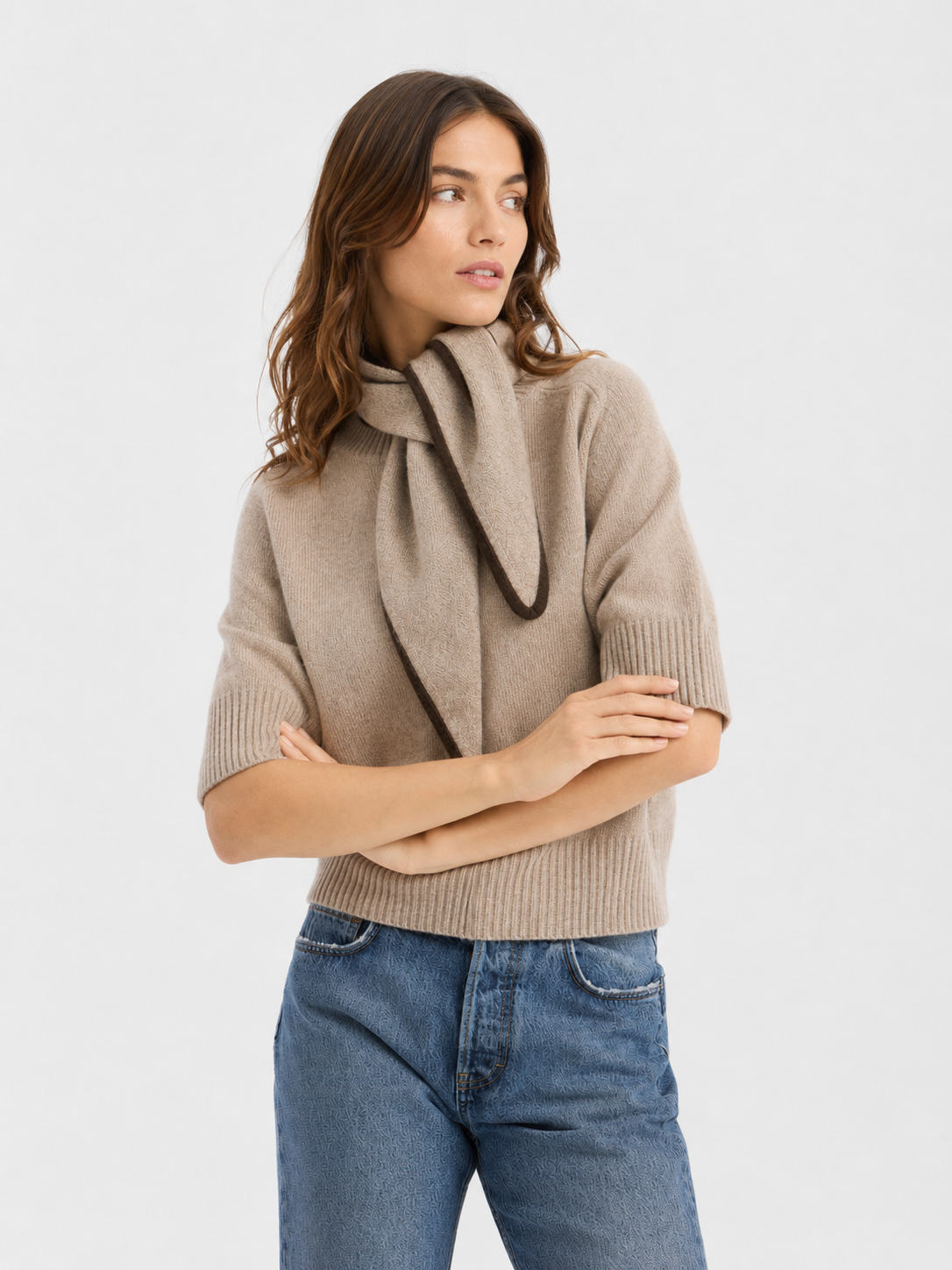 Eine Frau, die einen beigen Pullover, den Schal „Sophie Taupe“ und eine blaue Jeans trägt, steht mit verschränkten Armen vor einem schlichten weißen Hintergrund und blickt nach links.
