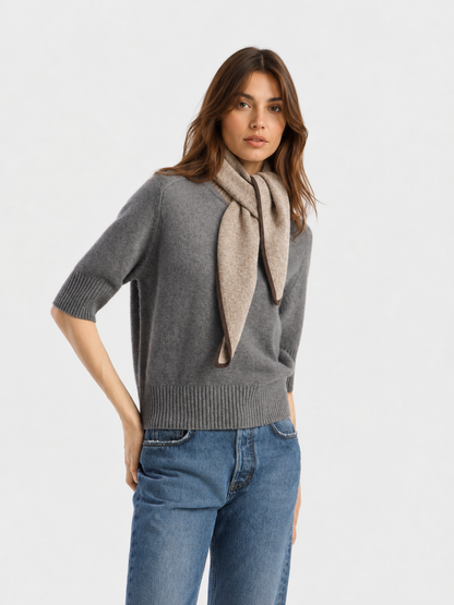 Eine Frau, die einen grauen Pullover mit halben Ärmeln und eine blaue Jeans trägt und den luxuriösen Schal „Sophie Taupe“ elegant um den Hals gebunden hat, steht vor einem schlichten weißen Hintergrund.