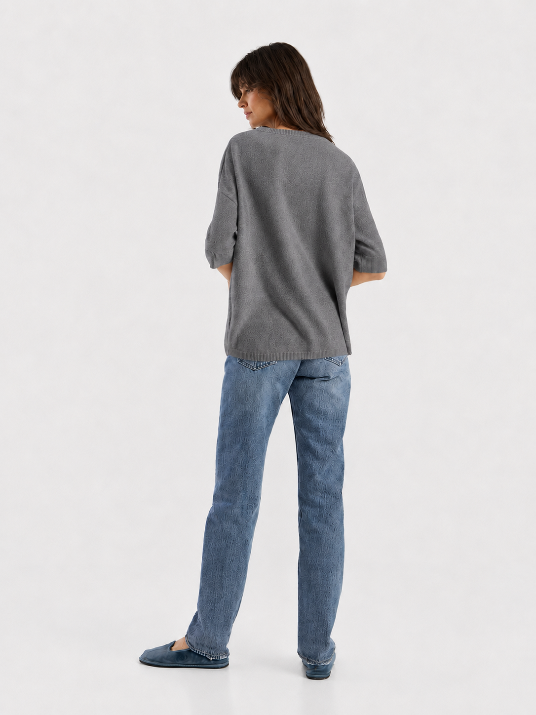 Eine Person steht mit dem Rücken zur Kamera und trägt ein locker sitzendes Strick-T-Shirt „Aurora Anthra“ in Grau, eine blaue Jeans und blaue Schuhe vor einem schlichten weißen Hintergrund.