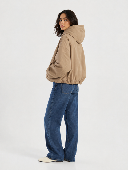 Eine Person steht seitlich vor einem schlichten weißen Hintergrund und trägt die „Jacket Savanna“, eine leichte Jacke mit Kapuze in Beige, dazu eine blaue Jeans und weiße Schuhe.