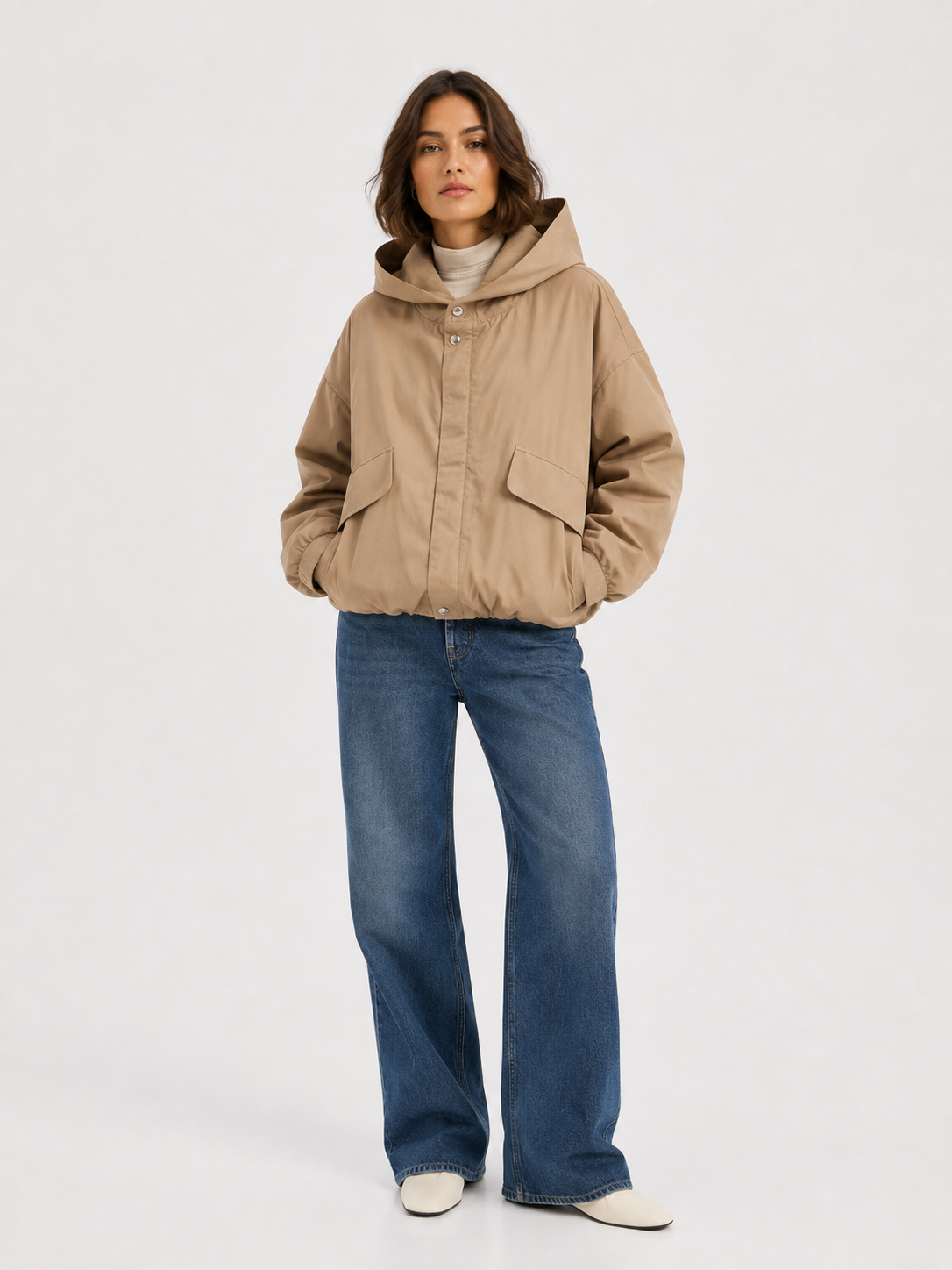Vor einem hellgrauen Hintergrund steht eine Frau, die die „Jacket Savanna“ trägt – eine beige Übergangsjacke für Damen mit übergroßer Kapuze, Druckknöpfen und Pattentaschen –, kombiniert mit einer blau-weit geschnittenen Jeans und weißen Schuhen.