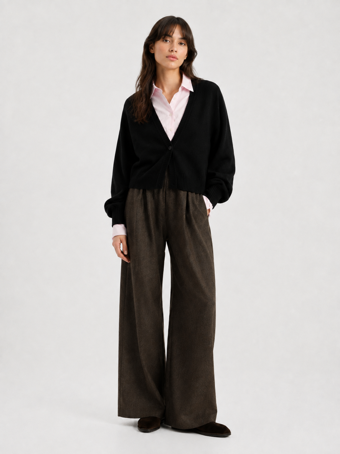 Vor einem schlichten Hintergrund steht eine Frau mit langen dunklen Haaren und ausdruckslosem Gesicht. Sie trägt die luxuriöse Antonia Short Cardigan Schwarz über einem hellrosa Hemd mit Kragen, dazu eine braune Hose mit weitem Bein und dunkle Schuhe.