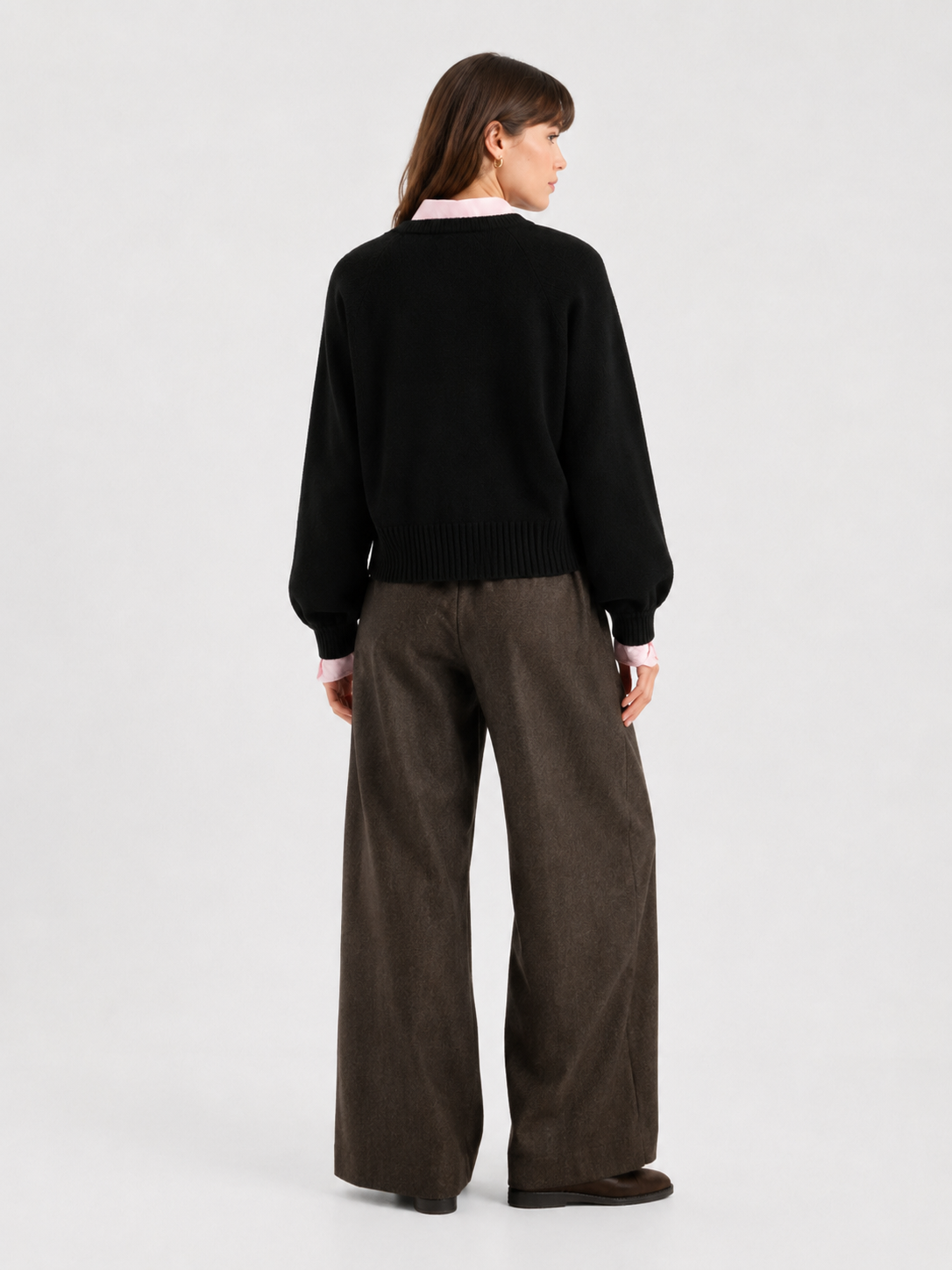 Vor einem schlichten, hellen Hintergrund steht eine Frau mit dem Rücken zur Kamera. Sie trägt den „Antonia Short Cardigan Schwarz“ über einem hellrosa Hemd, eine braune Hose mit weitem Bein und braune Schuhe.