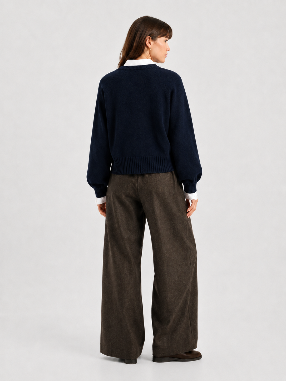 Eine Frau steht mit dem Rücken zur Kamera und trägt den Pullover „Charly Dunkelblau“ über einem weißen Hemd, eine braune Hose mit weitem Bein und braune Schuhe vor einem schlichten, hellen Hintergrund.