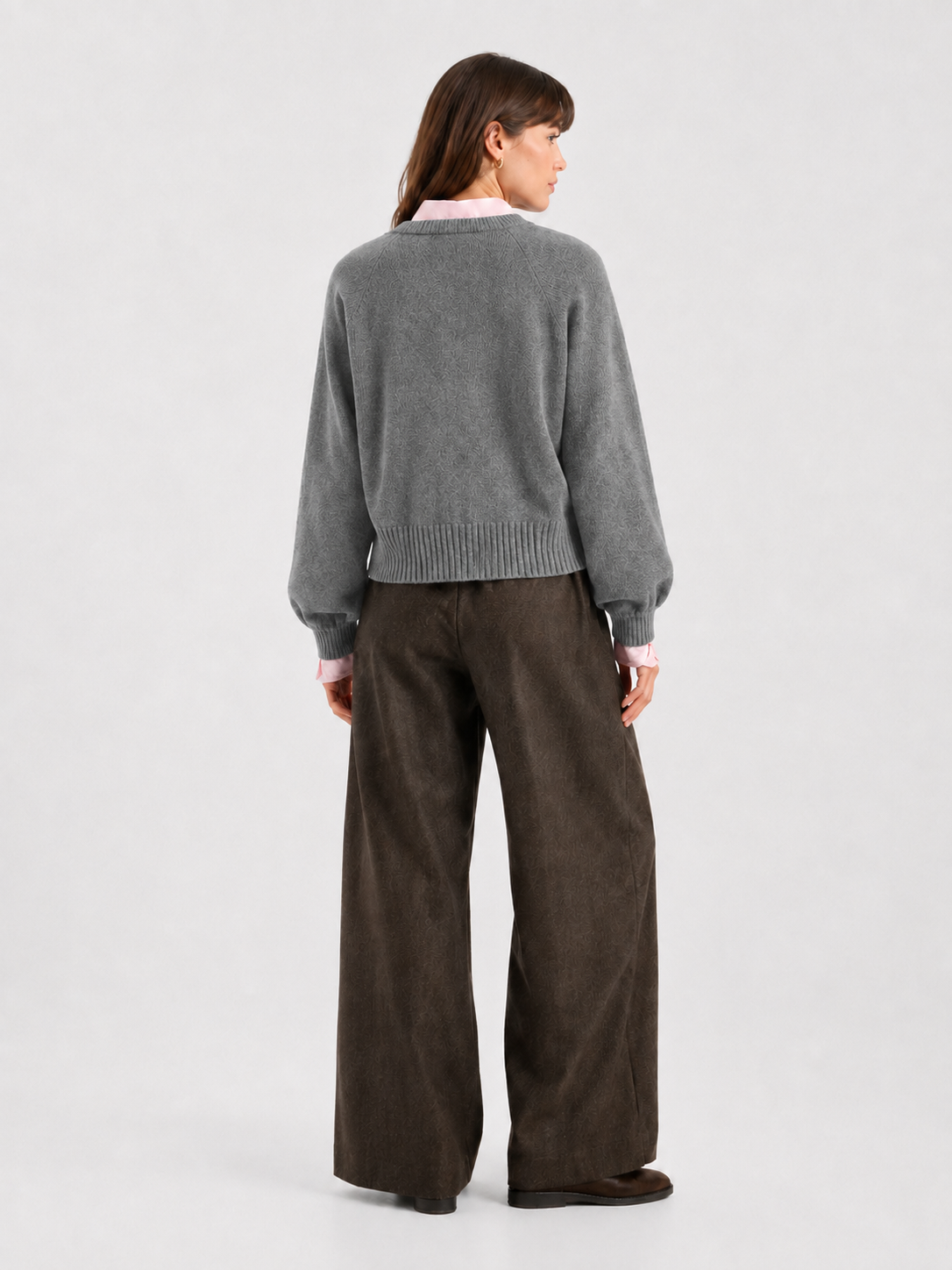 Eine Frau steht mit dem Rücken zur Kamera vor einem schlichten, hellen Hintergrund und trägt die kurze Strickjacke „Antonia“ in Grau über einem hellrosa Hemd mit Kragen und einer weiten braunen Hose.