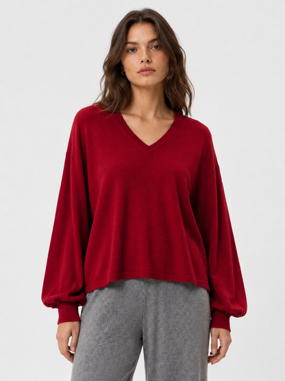 Eine Frau trägt den Pullover „Antonia V-Neck Rot“ mit lockerer Passform und langen Ärmeln sowie eine graue Hose und steht vor einem schlichten, hellgrauen Hintergrund.