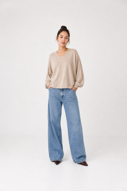 Eine Person steht vor einem schlichten Hintergrund und trägt den Pullover „Antonia V-Neck Powder“, eine blaue Jeans mit weitem Bein und braune spitze Schuhe; die Hände hat sie in den Taschen, und ihr Haar ist zu einem Dutt zusammengebunden.