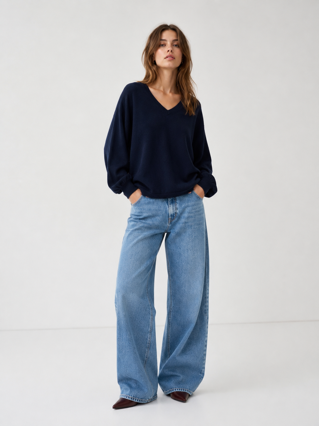 Eine Frau steht vor einem hellen Hintergrund und trägt den Pullover „Antonia V-Neck Dunkelblau“, dazu eine hellblaue Jeans mit weitem Bein und braune Schuhe; ihre Hände hat sie in die Taschen gesteckt.