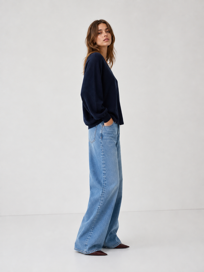 Eine Frau steht im Profil vor einem schlichten Hintergrund und trägt den Pullover „Antonia V-Neck Dunkelblau“ – einen locker sitzenden marineblauen Pullover – sowie eine blaue Jeans mit weitem Bein, wobei sie die Hände in die Taschen gesteckt hat. Ihr langes, welliges Haar umrahmt ihr Gesicht, während sie leicht in Richtung Kamera blickt.