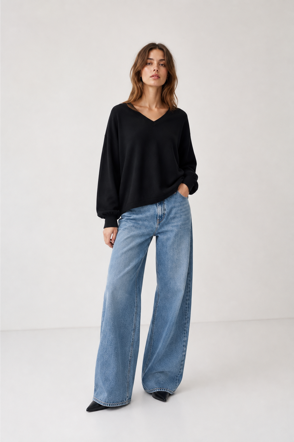 Eine Frau steht vor einem hellen Hintergrund und trägt den Pullover „Antonia V-Neck Schwarz“ sowie eine blaue Jeans mit weitem Bein. Eine Hand steckt in ihrer Hosentasche, während sie selbstbewusst in die Kamera blickt.