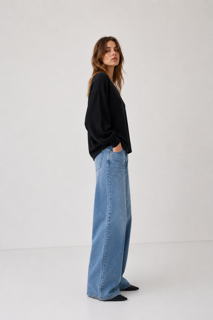 Eine Frau steht seitlich mit den Händen in den Taschen und trägt den Pullover „Antonia V-Neck Schwarz“ sowie eine blaue Jeans mit weitem Bein vor einem schlichten, hellen Hintergrund.