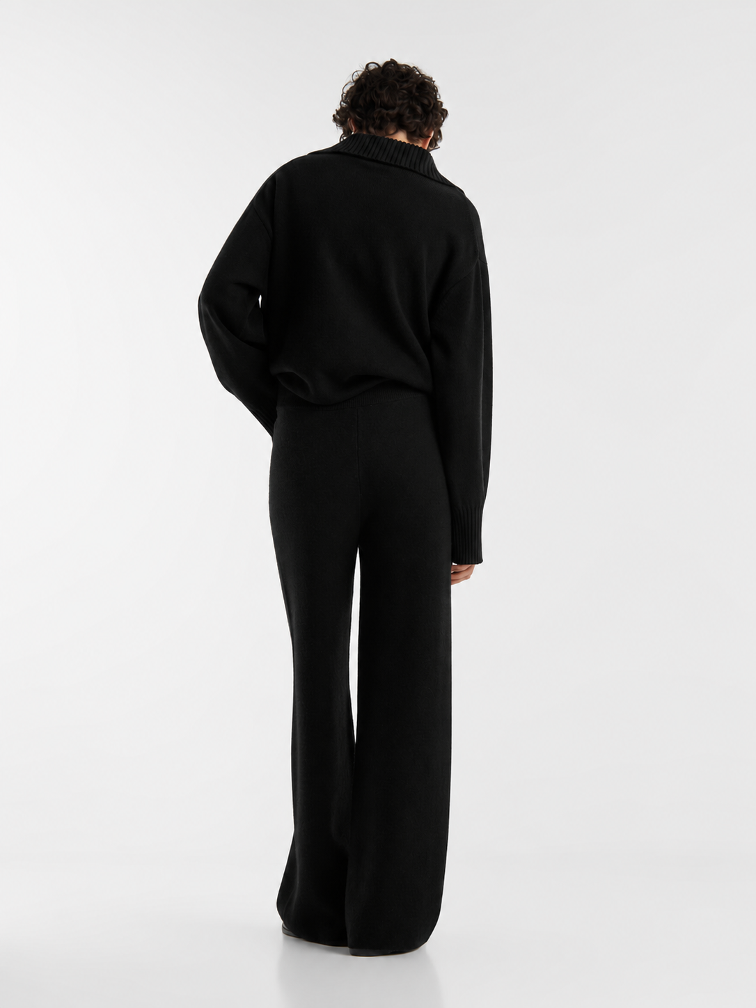 Eine Person mit kurzen Locken steht mit dem Rücken zur Kamera und trägt die Merino Hose Schwarz in Kombination mit einem schwarzen Strickpullover. Der minimalistische, einfarbige Look wird vor einem schlichten weißen Hintergrund in Szene gesetzt.