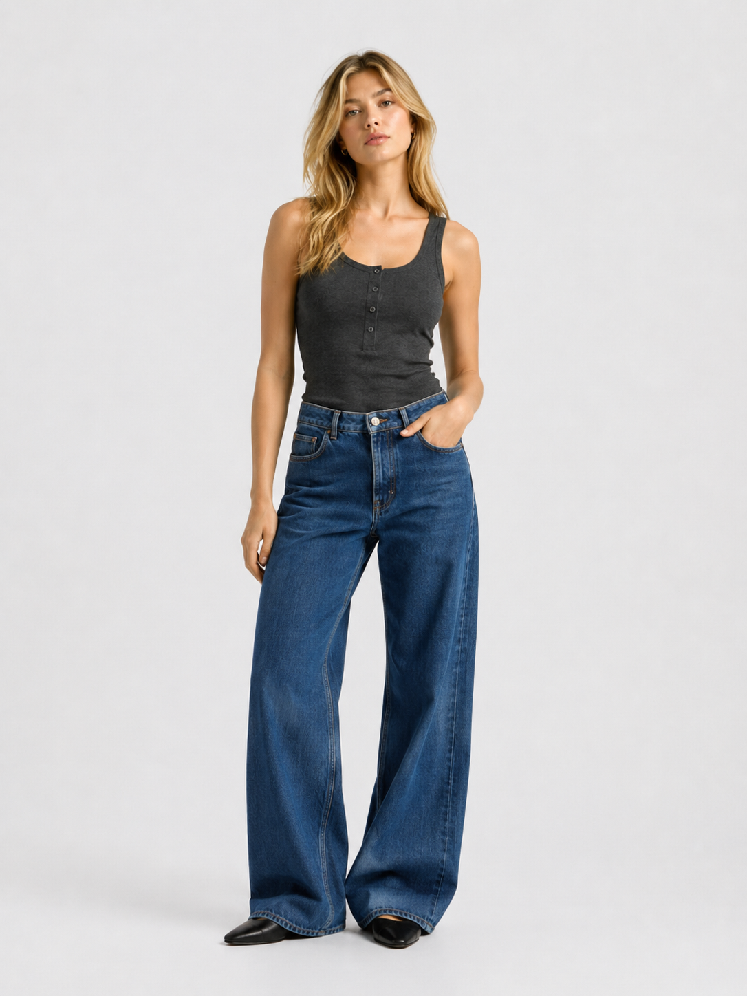 Eine Frau, die das figurbetonte Tanktop „Anthrazit“ und eine blaue Jeans mit weitem Bein trägt, steht mit einer Hand in der Hosentasche vor einem schlichten, hellen Hintergrund.