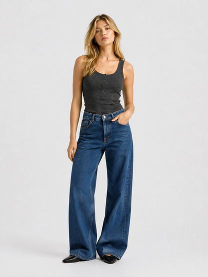Eine Frau, die das figurbetonte Tanktop „Anthrazit“ und eine blaue Jeans mit weitem Bein trägt, steht mit einer Hand in der Hosentasche vor einem schlichten, hellen Hintergrund.