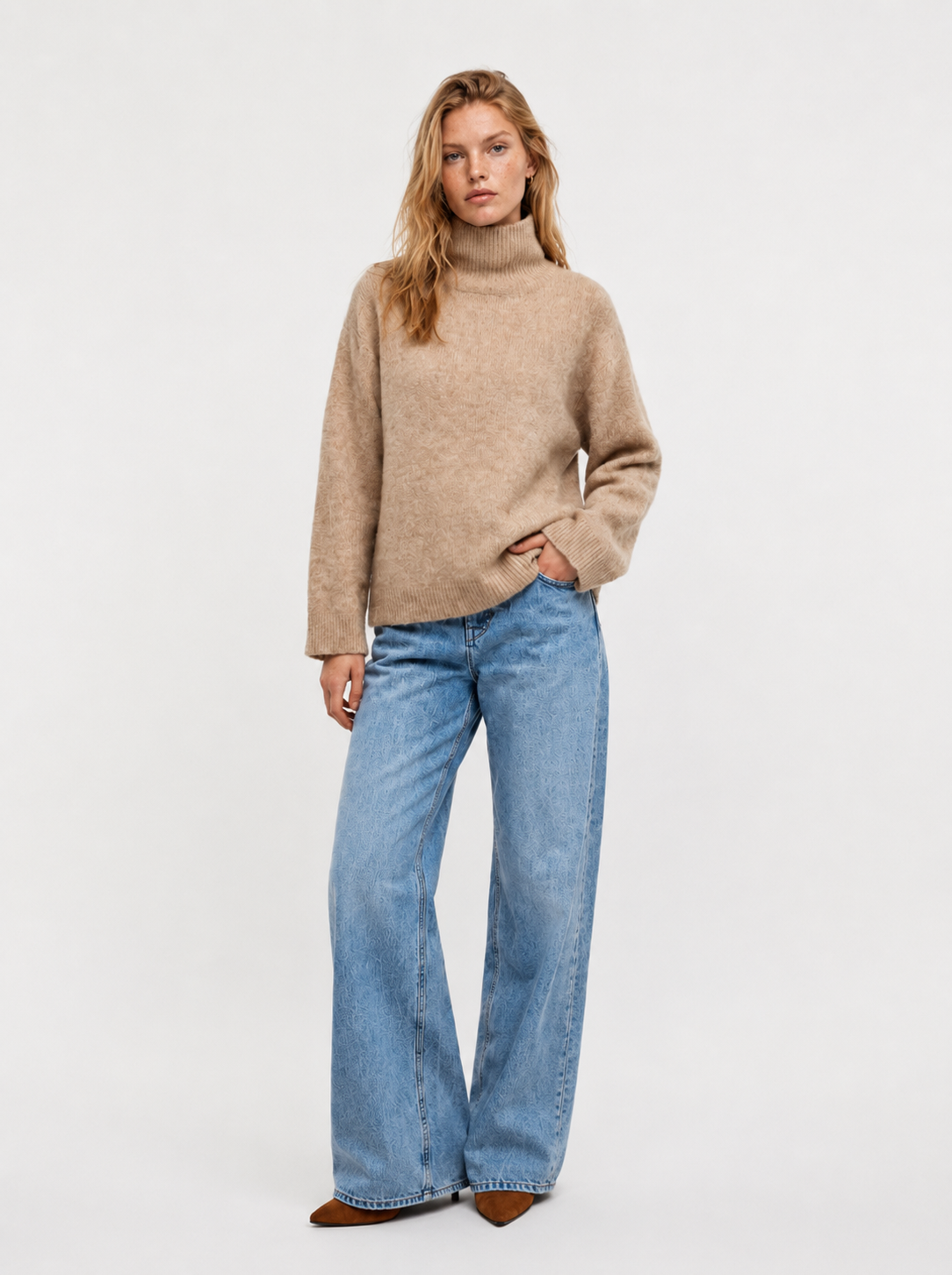 Eine Person steht vor einem schlichten Hintergrund und trägt den Pullover „Bear Turtle Vol. 2 Camel“, eine blaue Jeans mit weitem Bein und braune spitze Schuhe. Eine Hand steckt in der Tasche, und ihr Gesichtsausdruck ist neutral.