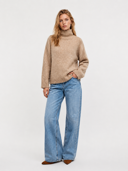 Eine Person steht vor einem schlichten Hintergrund und trägt den Pullover „Bear Turtle Vol. 2 Camel“, eine blaue Jeans mit weitem Bein und braune spitze Schuhe. Eine Hand steckt in der Tasche, und ihr Gesichtsausdruck ist neutral.