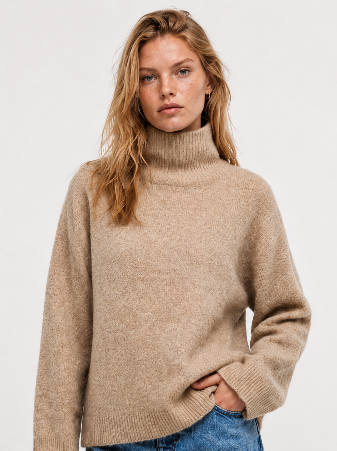 Eine junge Frau mit langen, welligen blonden Haaren trägt den „Bear Turtle Vol. 2 Camel“, einen luxuriösen beigen Rollkragenpullover, dazu eine blaue Jeans. Sie steht vor einem schlichten, hellen Hintergrund und blickt mit entspanntem Gesichtsausdruck direkt in die Kamera.