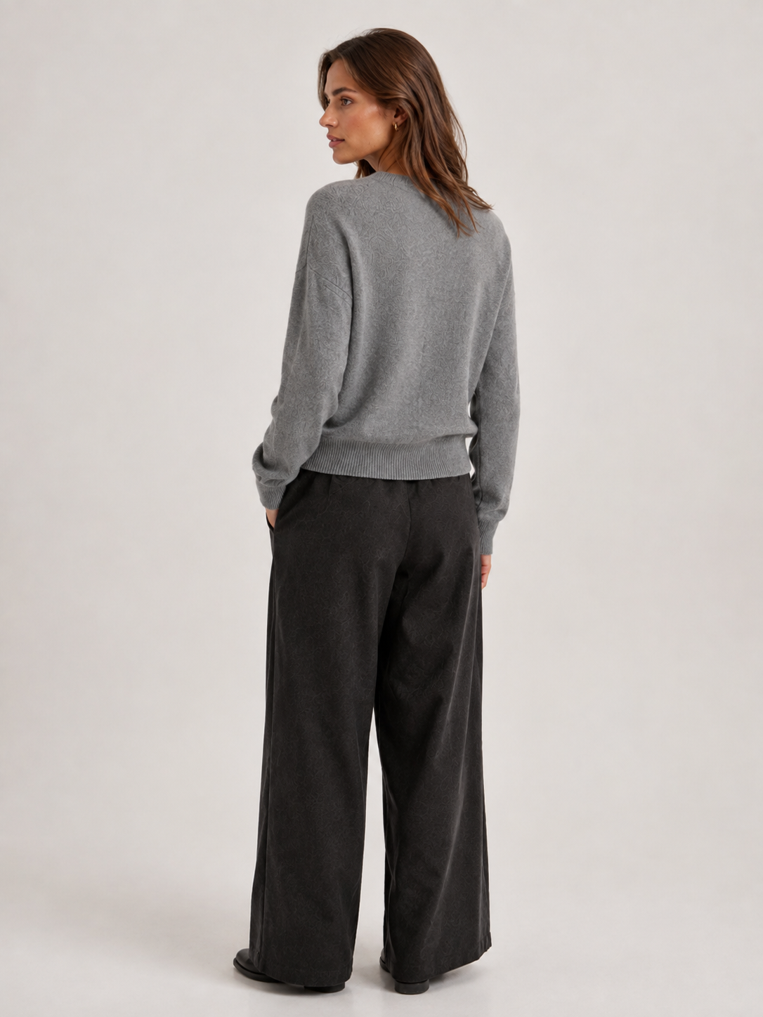 Eine Frau steht mit dem Rücken zur Kamera und trägt den Pullover „Tony Rundhals Grau“ aus einer weichen Merino-Kaschmir-Mischung sowie eine schwarze Hose mit weitem Bein. Sie steht leicht seitlich vor einem schlichten, hellen Hintergrund.