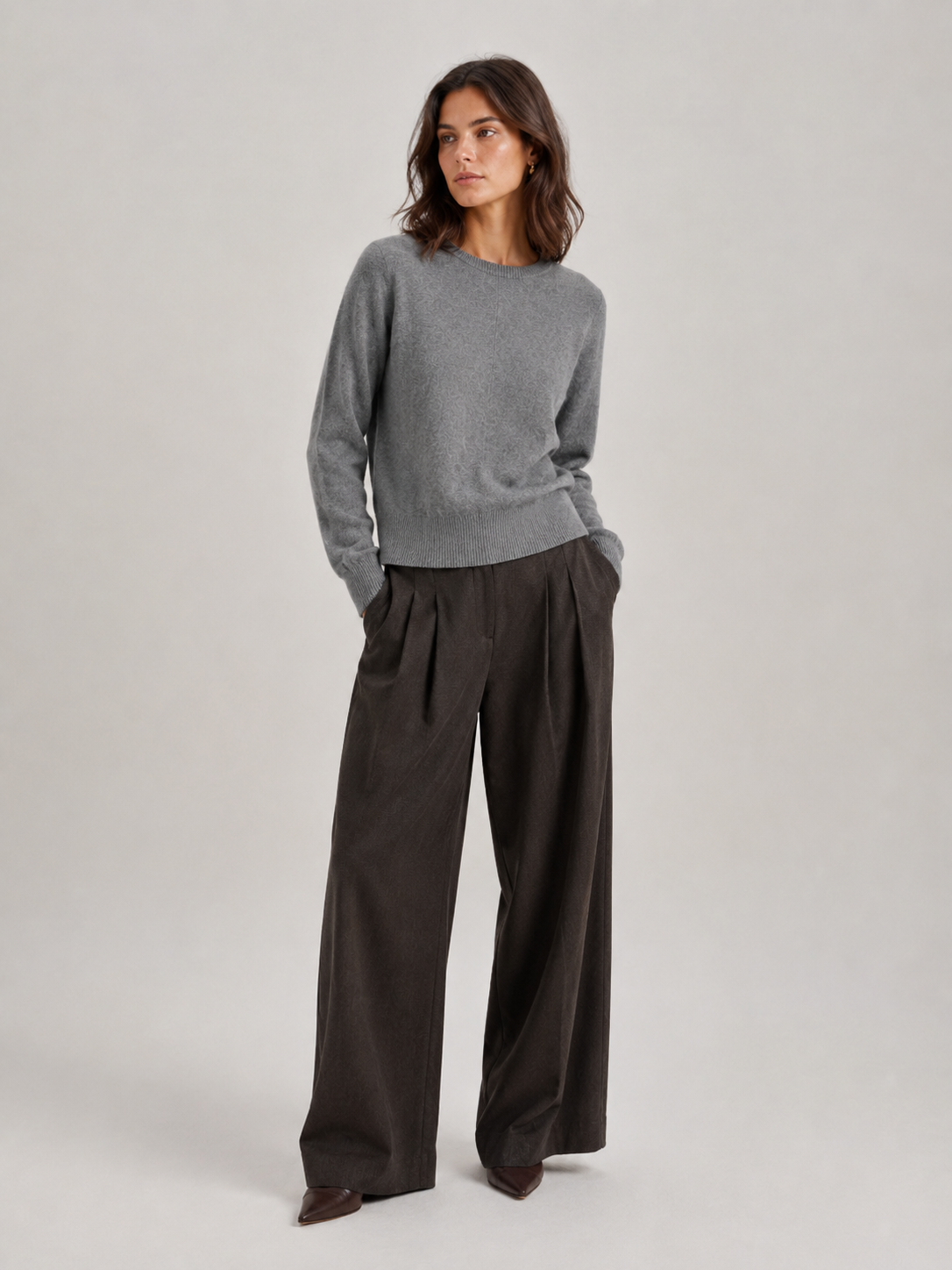 Eine Frau, die den Pullover „Tony Rundhals Grau“ und eine dunkelbraune Hose mit weitem Bein trägt, steht mit den Händen in den Taschen und blickt seitlich in die Kamera, vor einem schlichten, hellen Hintergrund.