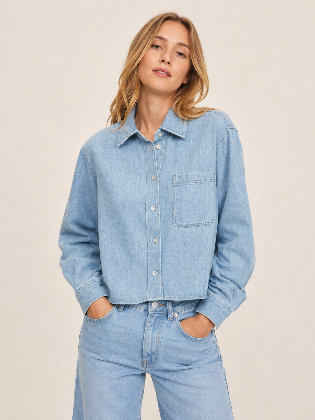 Eine Person mit langen, hellbraunen Haaren trägt die „Blouse Denim Cropped“ – ein hellblaues Jeanshemd mit Knöpfen und Brusttasche –, dazu eine passende Jeans mit hoher Taille, und steht vor einem schlichten, hellen Hintergrund.